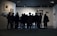 A group of people stands closely together in front of a well-lit display in an art gallery, observing the artwork. The gallery walls are adorned with several framed pieces, creating a contemplative atmosphere.