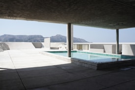 A modern architectural structure featuring a small swimming pool on a spacious terrace. The pool is surrounded by concrete surfaces and pillars, with a view of distant mountains under a clear sky.