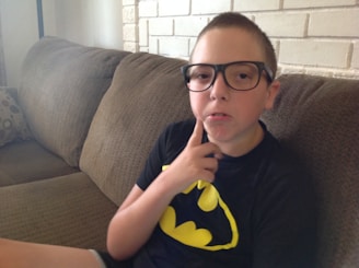 A young boy wearing large glasses and a Batman t-shirt is sitting on a brown couch. He holds his hand to his chin, suggesting a thoughtful pose. The background features a white brick wall.