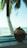 A fresh coconut with the top sliced open sits on a wooden table, with the backdrop of a serene ocean view and a palm tree. The scene suggests a tropical setting with a relaxed atmosphere.