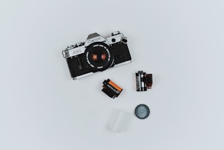 A vintage Canon AE-1 camera is placed on a plain white background. Surrounding the camera are various photography accessories including two film canisters, a film roll, a lens cap, and a small plastic container.