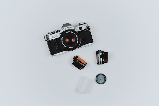A vintage Canon AE-1 camera is placed on a plain white background. Surrounding the camera are various photography accessories including two film canisters, a film roll, a lens cap, and a small plastic container.