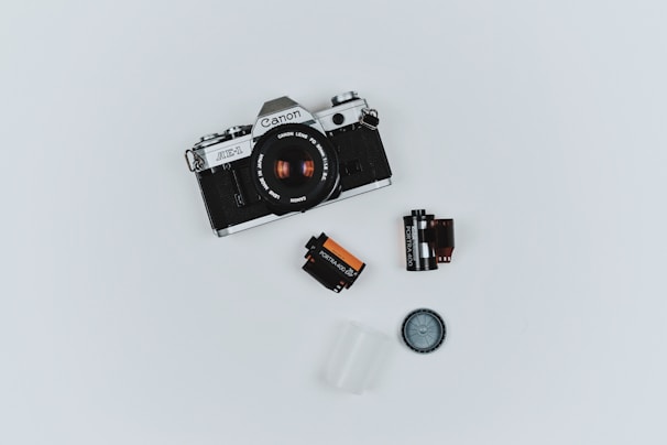 A vintage Canon AE-1 camera is placed on a plain white background. Surrounding the camera are various photography accessories including two film canisters, a film roll, a lens cap, and a small plastic container.