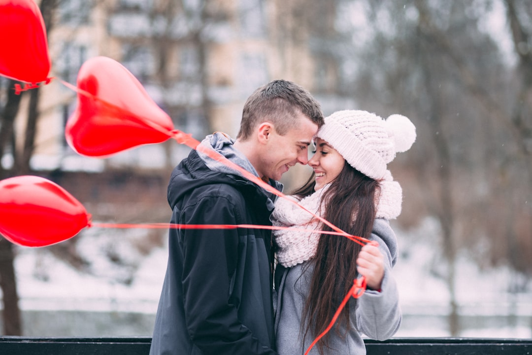 A man and woman smiling while touching face to face, as the girl holds red heart shaped balloons.