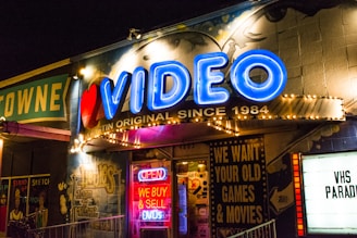 A brightly lit storefront with a neon sign reading 'VIDEO' in blue, accompanied by a red heart. The facade is adorned with various retro-style signs, including one offering to buy and sell DVDs. The atmosphere suggests a vintage video rental store, with additional signage advertising the purchase of old games and movies. The storefront has a lively and nostalgic appearance, with graffiti-style artwork on the walls and colorful lighting.