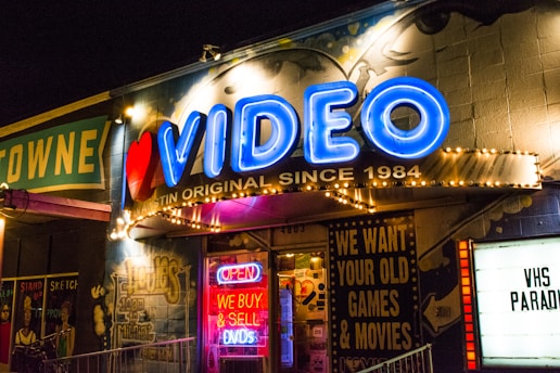A brightly lit storefront with a neon sign reading 'VIDEO' in blue, accompanied by a red heart. The facade is adorned with various retro-style signs, including one offering to buy and sell DVDs. The atmosphere suggests a vintage video rental store, with additional signage advertising the purchase of old games and movies. The storefront has a lively and nostalgic appearance, with graffiti-style artwork on the walls and colorful lighting.