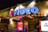 A brightly lit storefront with a neon sign reading 'VIDEO' in blue, accompanied by a red heart. The facade is adorned with various retro-style signs, including one offering to buy and sell DVDs. The atmosphere suggests a vintage video rental store, with additional signage advertising the purchase of old games and movies. The storefront has a lively and nostalgic appearance, with graffiti-style artwork on the walls and colorful lighting.