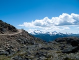 Three hikers stand on a rocky mountain trail with a panoramic view of snow-capped mountains and a bright blue sky scattered with fluffy clouds. The rough texture of the rocks and the expansive landscape create a sense of adventure and exploration.