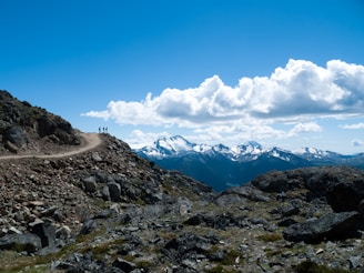 Three hikers stand on a rocky mountain trail with a panoramic view of snow-capped mountains and a bright blue sky scattered with fluffy clouds. The rough texture of the rocks and the expansive landscape create a sense of adventure and exploration.