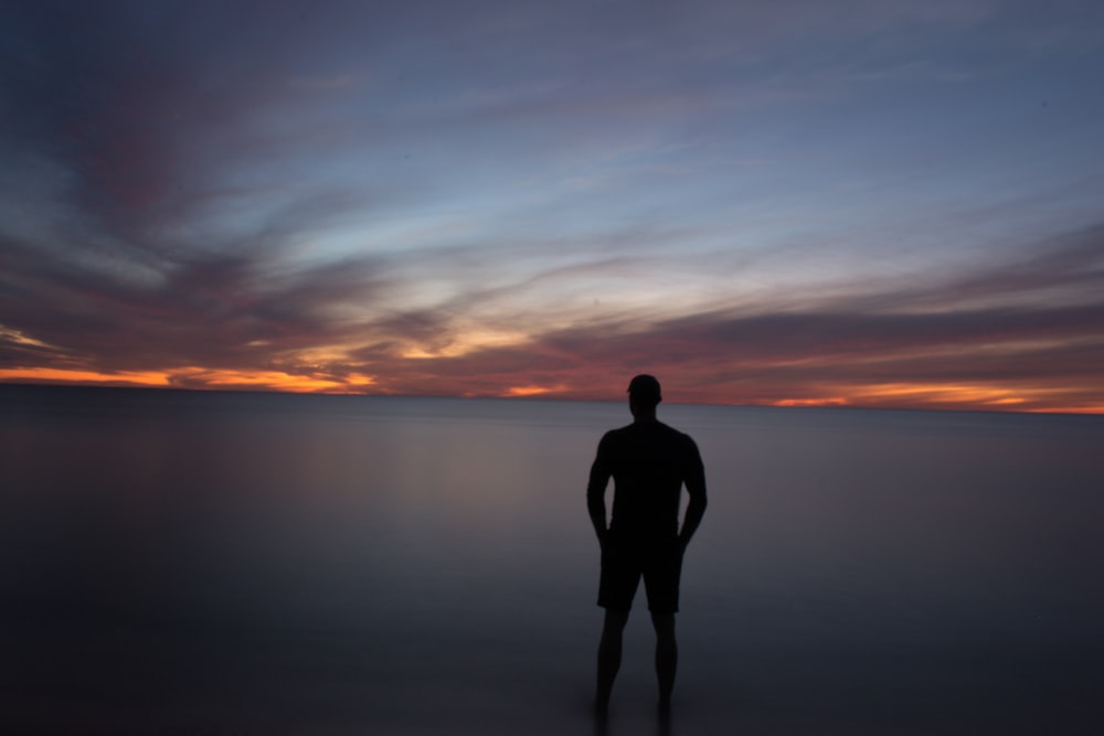 silhouette of person standing near calm body of water during golden hour