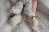 A baby's legs are shown wearing soft, fluffy pants and knitted cream booties with brown ribbon ties, all set against a plush white blanket.