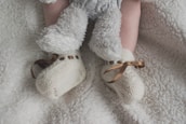 A baby's legs are shown wearing soft, fluffy pants and knitted cream booties with brown ribbon ties, all set against a plush white blanket.