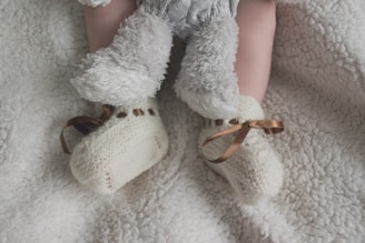 A baby's legs are shown wearing soft, fluffy pants and knitted cream booties with brown ribbon ties, all set against a plush white blanket.