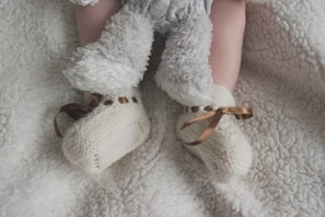 A baby's legs are shown wearing soft, fluffy pants and knitted cream booties with brown ribbon ties, all set against a plush white blanket.