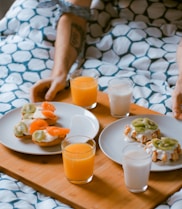 person serving pastries on white ceramic plates with fruit juice glasses on wooden tray on top of bed