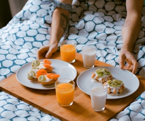 person serving pastries on white ceramic plates with fruit juice glasses on wooden tray on top of bed