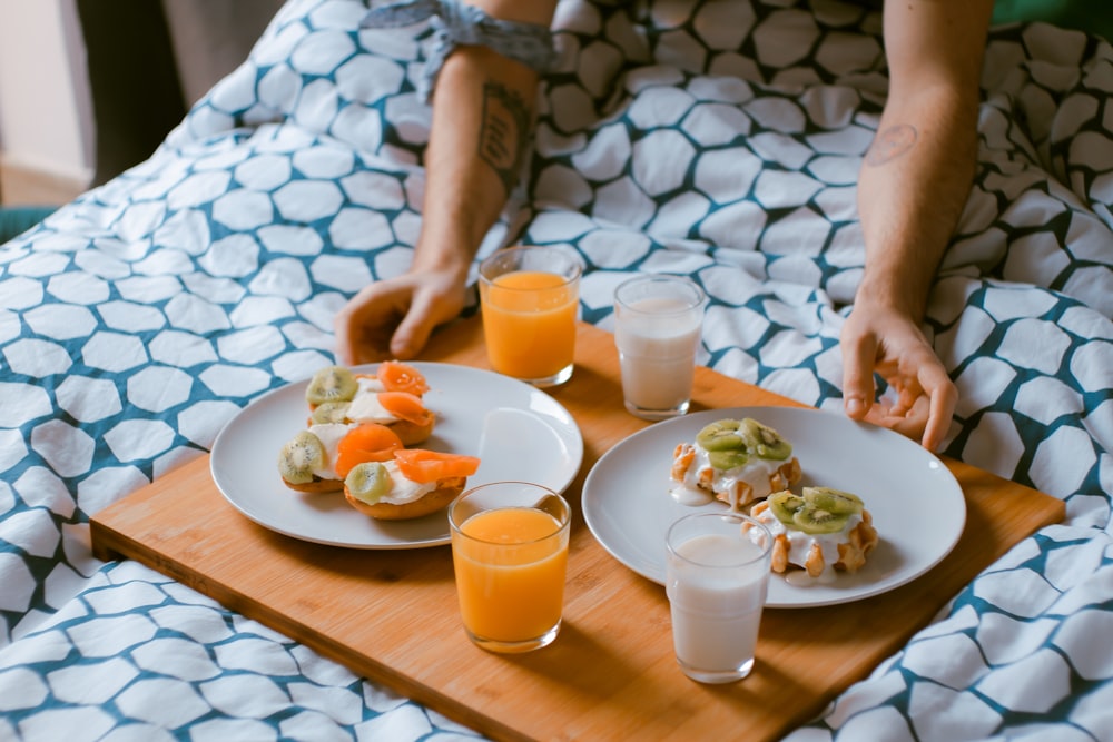 침대 위의 나무 쟁반에 과일 주스 잔이 있는 흰색 세라믹 접시에 페이스트리를 제공하는 사람