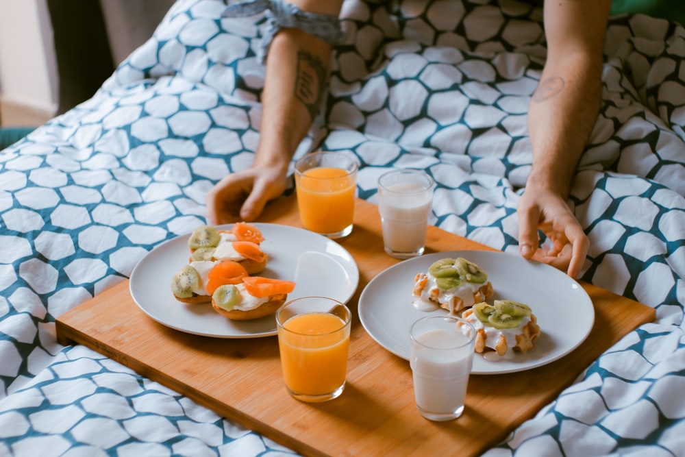 person serving pastries on white ceramic plates with fruit juice glasses on wooden tray on top of bed