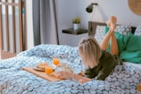 A person is lying on their stomach on a bed covered with geometric patterned bedding. They are looking at a breakfast tray that holds a glass of orange juice, a plate of food with vegetables, and a cup. The room has modern decor with a small plant, a wall-mounted lamp, and curtains.