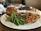 A gourmet plate with a hamburger featuring a glossy, toasted bun, accompanied by fresh mixed greens and crunchy onion rings. A small dish with a red dipping sauce is also present, enhancing the sophisticated dining presentation.