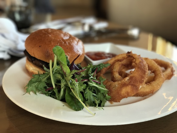 A gourmet plate with a hamburger featuring a glossy, toasted bun, accompanied by fresh mixed greens and crunchy onion rings. A small dish with a red dipping sauce is also present, enhancing the sophisticated dining presentation.