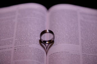 An open book with a ring standing upright on its pages, casting a shadow in the shape of a heart. The focus is on the center of the ring and the text around it is blurred.