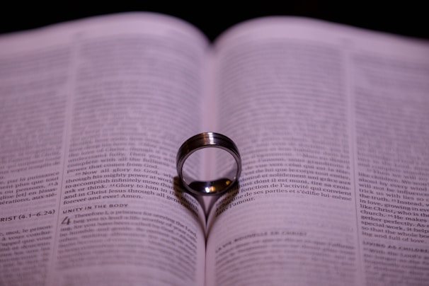 An open book with a ring standing upright on its pages, casting a shadow in the shape of a heart. The focus is on the center of the ring and the text around it is blurred.