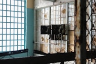 An old, rusted electrical panel is mounted on a wall with broken windows. The windows have a grid pattern, and some panes are missing or cracked. Rust and decay are evident on the metal structures, indicating neglect and abandonment. Sunlight filters in, casting shadows and highlighting the deteriorated state of the room.