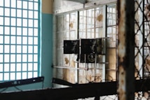 An old, rusted electrical panel is mounted on a wall with broken windows. The windows have a grid pattern, and some panes are missing or cracked. Rust and decay are evident on the metal structures, indicating neglect and abandonment. Sunlight filters in, casting shadows and highlighting the deteriorated state of the room.