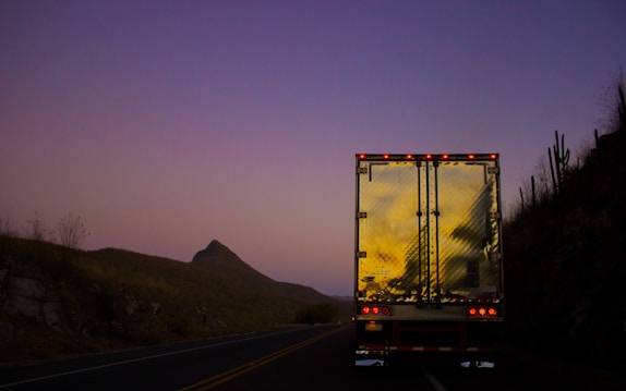 A large truck with a reflective metallic back is driving down a road during sunset or dusk. The sky is a gradient of soft pink and purple hues, and the truck's reflection captures the warm glow of the setting sun. Surrounding the road are hills and sparse vegetation, including some cacti, under the dim light.
