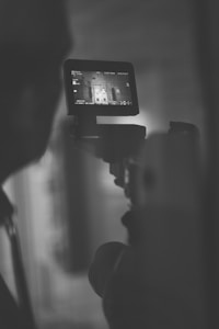 A person is holding a camera or camcorder, capturing footage. The focus is on the screen of the device, which displays a monochrome image of an interior scene. The overall ambiance is dark, with the silhouette of the camera operator visible.
