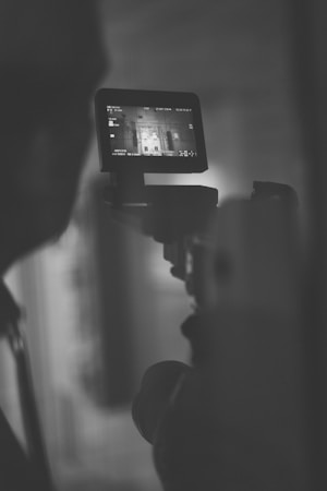 A person is holding a camera or camcorder, capturing footage. The focus is on the screen of the device, which displays a monochrome image of an interior scene. The overall ambiance is dark, with the silhouette of the camera operator visible.