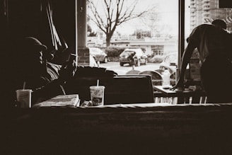 A dimly lit indoor scene features two people in a cafe setting; one is sitting and appears to be looking at a phone, while the other is standing at a table with items like drinks and pizza boxes visible. Through the window, an urban street with parked cars and bare trees is visible, suggesting a wintry or early spring atmosphere.
