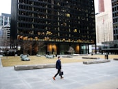 A man in formal attire, holding a briefcase, is walking across a large urban plaza. The plaza features modern architectural elements including a black-gridded high-rise building. There is a flagpole with a red and white flag, bare trees, and several large sculptures of reclining cows situated on patches of grass.