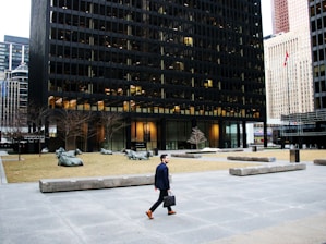 A man in formal attire, holding a briefcase, is walking across a large urban plaza. The plaza features modern architectural elements including a black-gridded high-rise building. There is a flagpole with a red and white flag, bare trees, and several large sculptures of reclining cows situated on patches of grass.