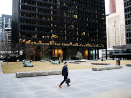 A man in formal attire, holding a briefcase, is walking across a large urban plaza. The plaza features modern architectural elements including a black-gridded high-rise building. There is a flagpole with a red and white flag, bare trees, and several large sculptures of reclining cows situated on patches of grass.