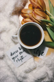 A cozy setup featuring a cup of black coffee, a small bouquet of orange tulips, and a fluffy white fabric. Two cards with handwritten messages, 'dance to your own rhythm' and 'love', are placed near the coffee. The scene is set on a wooden surface, creating an inviting and warm ambiance.