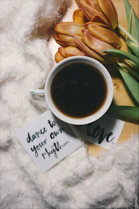 A cozy setup featuring a cup of black coffee, a small bouquet of orange tulips, and a fluffy white fabric. Two cards with handwritten messages, 'dance to your own rhythm' and 'love', are placed near the coffee. The scene is set on a wooden surface, creating an inviting and warm ambiance.