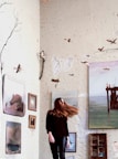 A woman stands in a room with textured, artistic walls adorned with paintings and small sculptures. Her hair is caught in mid-motion, adding a dynamic element. The walls have a dotted pattern, and there are branches attached, giving an organic feel. The paintings depict various landscapes and seascapes, creating an eclectic atmosphere.