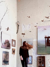 A woman stands in a room with textured, artistic walls adorned with paintings and small sculptures. Her hair is caught in mid-motion, adding a dynamic element. The walls have a dotted pattern, and there are branches attached, giving an organic feel. The paintings depict various landscapes and seascapes, creating an eclectic atmosphere.