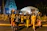 A group of people dressed in colorful traditional attire are participating in a night procession. Some of them are carrying decorative objects on their heads, and they are adorned with floral garlands. The backdrop features an illuminated temple structure.