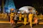 A group of people dressed in colorful traditional attire are participating in a night procession. Some of them are carrying decorative objects on their heads, and they are adorned with floral garlands. The backdrop features an illuminated temple structure.