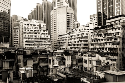 An urban landscape featuring a dense collection of high-rise and mid-rise buildings in various architectural styles. Some buildings appear older and slightly worn, with visible air conditioning units and balconies, while others are more modern with glass facades. The scene is predominantly monochrome, adding a vintage or timeless feel.