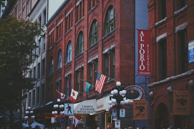 A bustling urban street scene features buildings with red brick facades and arched windows. Signs indicate various businesses, including a restaurant named Al Porto Ristorante Italiano. Several flags are visible, including the American and Canadian flags. Alongside signs for souvenirs and native crafts, there are numerous streetlights and hanging flower baskets. A tree is visible on the left, adding greenery to the urban landscape.