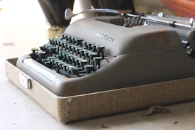 A vintage typewriter with a metal body and green keys sits inside an open, textured case. The machine shows signs of rust and wear, highlighting its age and frequent use.
