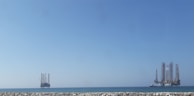 A seascape featuring two offshore oil rigs positioned in the calm sea. The larger rig is on the right, and a smaller one is on the left, both set against a clear blue sky. In the foreground, a stone jetty separates the water from the viewer.