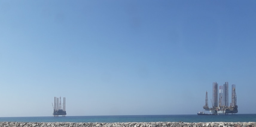 A seascape featuring two offshore oil rigs positioned in the calm sea. The larger rig is on the right, and a smaller one is on the left, both set against a clear blue sky. In the foreground, a stone jetty separates the water from the viewer.