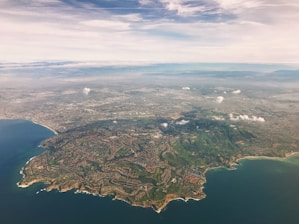 An aerial view of a wide coastal area with sprawling urban and suburban development. The coastline is bordered by clear blue water, and the land is lush with green hills and residential areas. Small patches of clouds are scattered in the sky, adding depth to the scene.