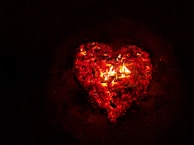 A glowing heart shape formed by embers and flames against a dark background. The heart is outlined in bright red and orange colors, giving it a vibrant and intense appearance.