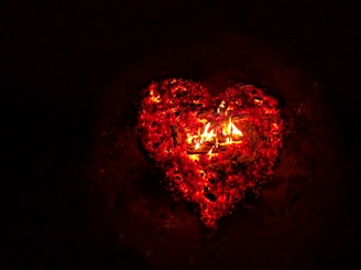 A glowing heart shape formed by embers and flames against a dark background. The heart is outlined in bright red and orange colors, giving it a vibrant and intense appearance.